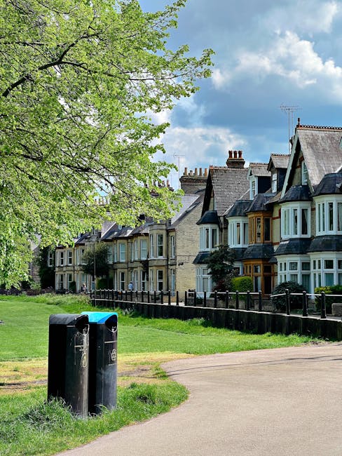 A quiet residential street scene showing a row of traditional Victorian terraced houses with bay windows, pitched slate roofs, and brick chimneys, situated behind a low black metal fence. In the foreground, a small patch of well-maintained green grass borders a light brown concrete pathway, with two black public litter bins positioned on the grass near the pathway's edge. A large leafy tree with bright green foliage extends over the scene from the left, casting shade over part of the area. The sky above is partly cloudy with patches of blue visible through scattered white clouds. The scene captures an ordinary suburban setting that may relate to private waste disposal or the need for local rubbish collection services, with the presence of waste bins indicating waste management practices in a typical residential neighbourhood, possibly serviced by independent rubbish removal providers like Rubbish Clearance Wimbledon.