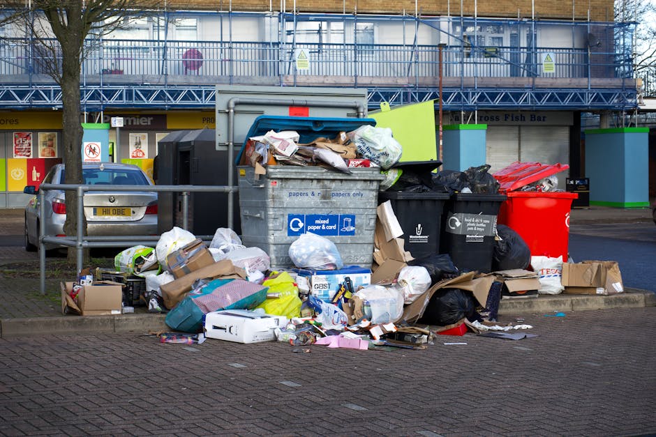 A large collection of overflowing rubbish bins and piles of waste around a designated disposal area on a paved urban sidewalk. The scene includes various types of waste, such as cardboard boxes, plastic bags, paper packaging, and mixed debris, some of which are spilling onto the ground. There are several bins of different colours, notably a grey bin labeled for mixed paper and card, a black bin, and a red bin, all filled with or surrounded by waste materials. An open green lid on the grey bin reveals crumpled papers and cardboard. Behind the waste area, a parked silver car is partially visible, with a traditional UK license plate. The background features a storefront with closed shutters, a bright yellow sign, and a multi-story building under construction or renovation, surrounded by scaffolding and safety netting. The lighting suggests an overcast day, creating a neutral, professional tone suitable for waste management or rubbish removal context.