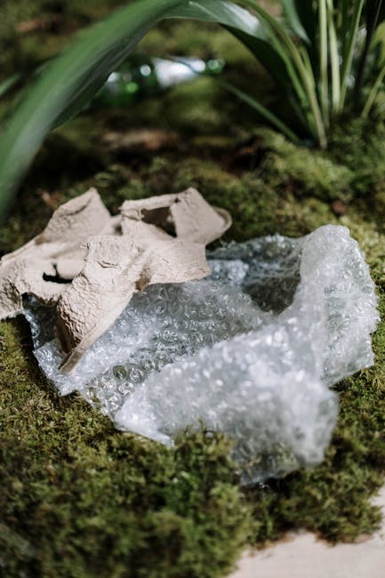 A close-up image shows broken ceramic fragments and a piece of clear bubble wrap resting on a moss-covered ground, surrounded by green foliage and plant leaves. The ceramic pieces appear to be light beige with a textured surface, possibly parts of an unused or discarded item, and are positioned towards the upper left of the image. The bubble wrap, located in the foreground, is transparent with small air-filled bubbles giving it a shiny, textured surface. The environment appears natural, with the moss providing a soft, uneven ground cover and the background featuring leafy plants with large, dark green, elongated leaves that arch slightly over the scene. The lighting is natural, diffused with no harsh shadows, creating a calm, outdoor atmosphere. This scene could suggest a setting where waste such as packaging materials and broken ceramics are temporarily placed before proper disposal, aligning with private or alternative rubbish collection methods where on-site clearance could be involved, as seen by the proximity of waste items within a natural environment.