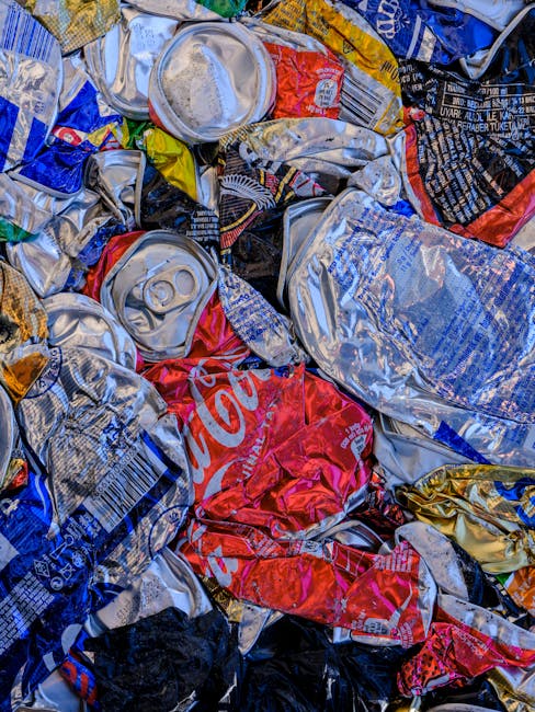 A close-up image of a pile of crushed aluminum and tin cans, including recognizable brands such as Coca-Cola, with metallic surfaces that are wrinkled and compressed. The cans are irregularly shaped, overlapping each other, and display a variety of bright colors such as red, blue, yellow, and silver. Some cans are stacked or leaning against each other in a disorderly manner, with visible pull tabs and partial labels. The background environment suggests they are outdoors or in a waste collection area, with no visible container or vehicle, indicating a scene of waste awaiting collection or recycling. The lighting highlights the reflective surfaces of the cans, emphasizing their textures and colors. This image relates to waste management and private rubbish disposal services provided by Rubbish Clearance Wimbledon, showcasing the type of recyclable materials handled during an alternative waste collection process, such as on-site clearance or waste recycling services.